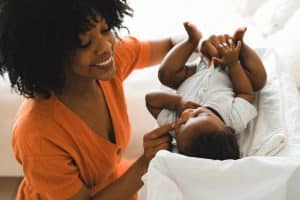 Mãe sorridente com vestido laranja interage carinhosamente com seu bebê deitado em uma superfície acolchoada, em um ambiente iluminado e acolhedor.